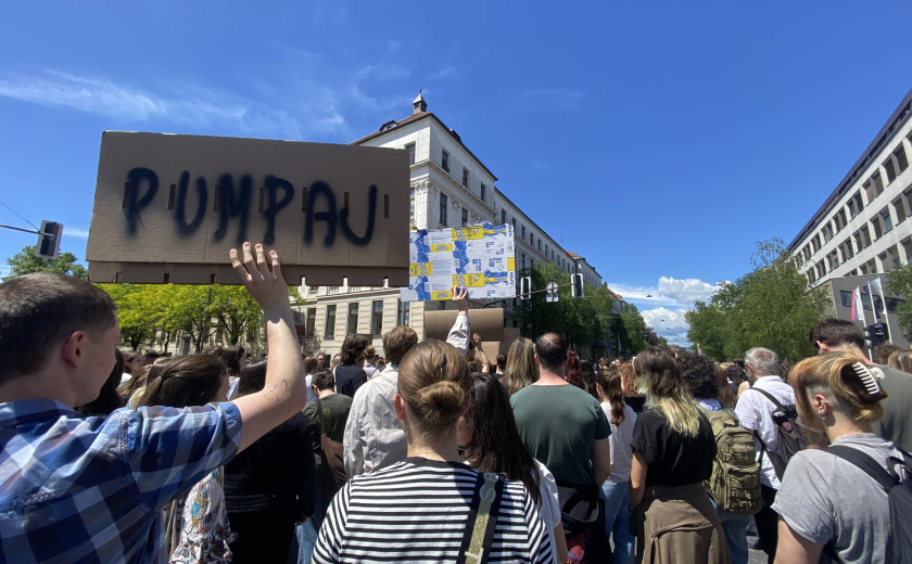 Fotografija večjega števila protestnikov, eden od njih drži kartonast transparent, na katerem z velikimi tiskanimi črkami piše pumpaj